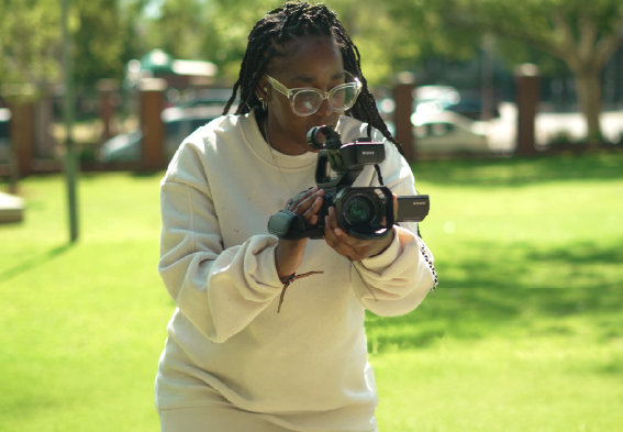 Individual holding a video camera while recording outdoors in a green space.