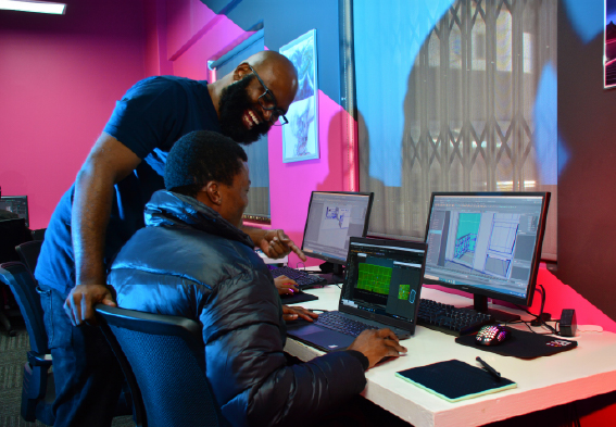Instructor assisting a student with computer programming at a workspace with dual monitors.